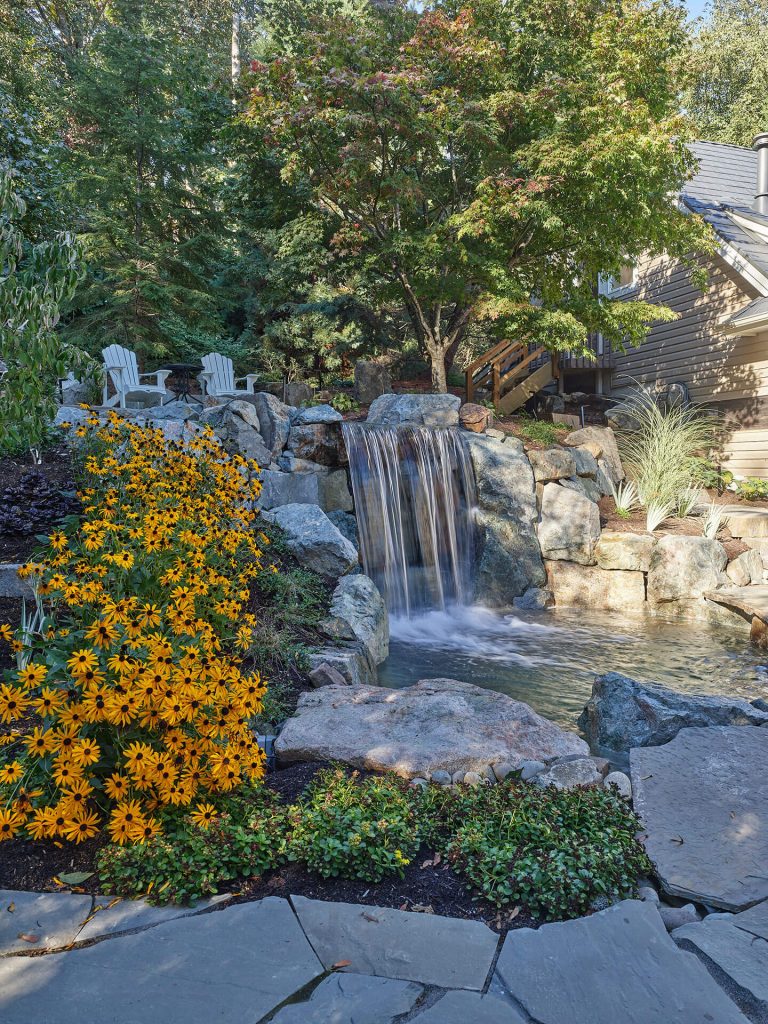 A waterfall pours into a rock-lined pool beneath mature trees with seating areas above.