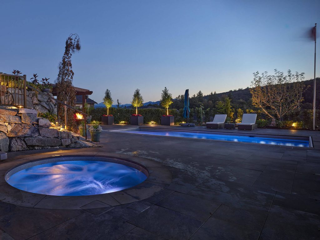 A glowing round spa beside a long lap pool, with soft landscape lighting and distant hillside views.