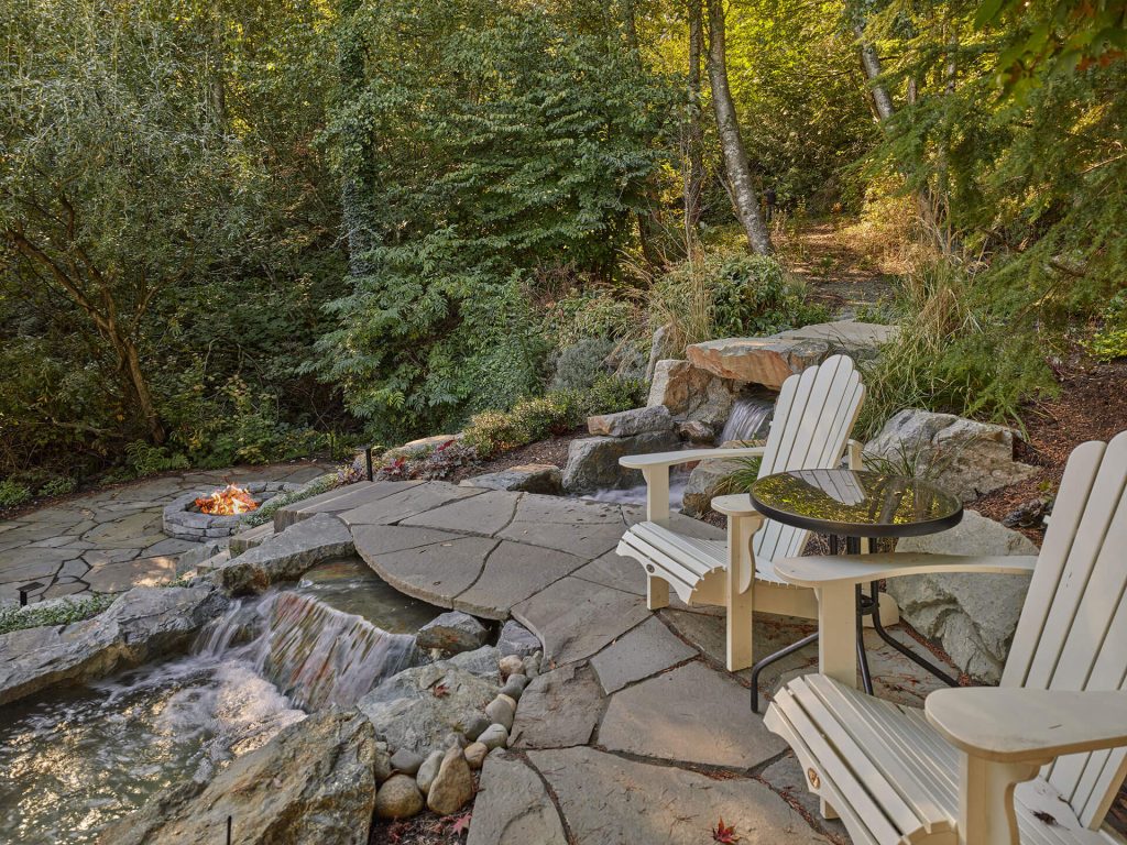Adirondack chairs overlook a stream and firepit tucked into a forested landscape with stone paths.
