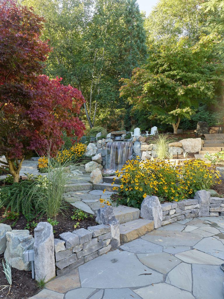 A stone pathway leads to a landscaped waterfall surrounded by bright flowers and mature trees.