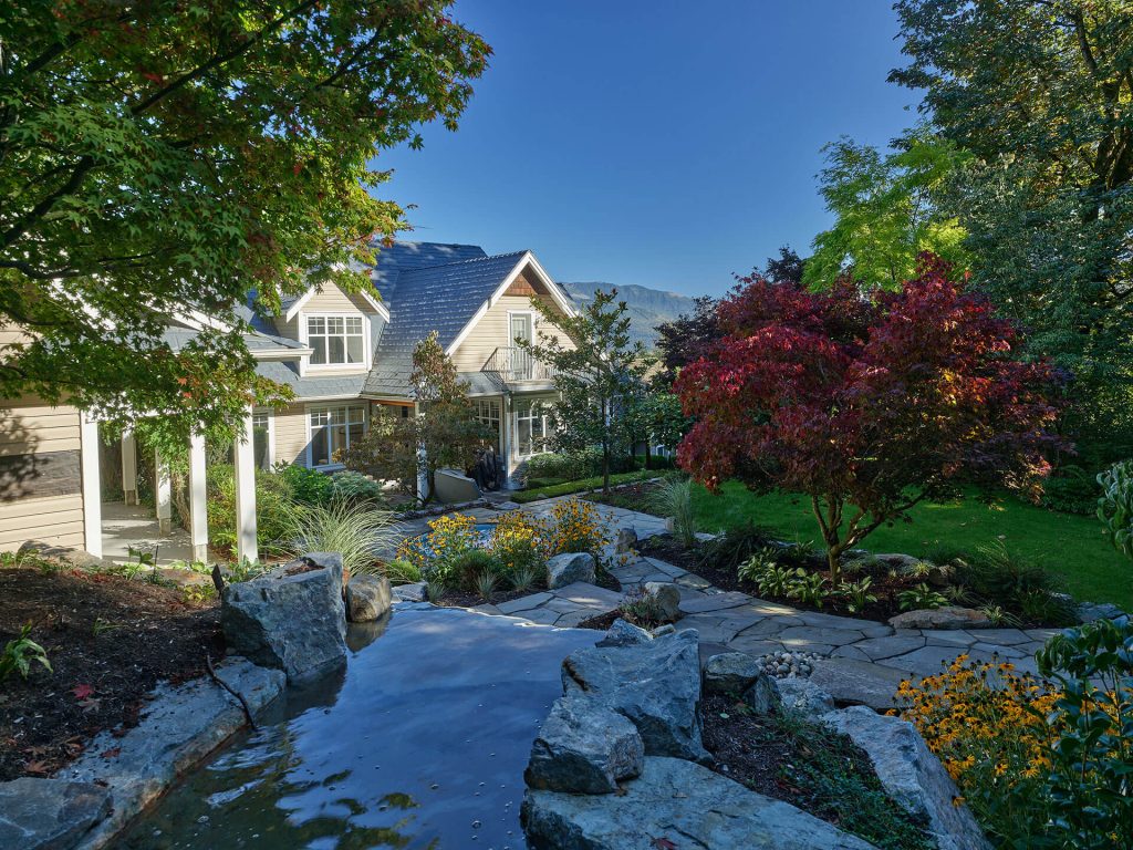 A landscaped stream flows through natural stone toward a home framed by colorful trees and mountain views.