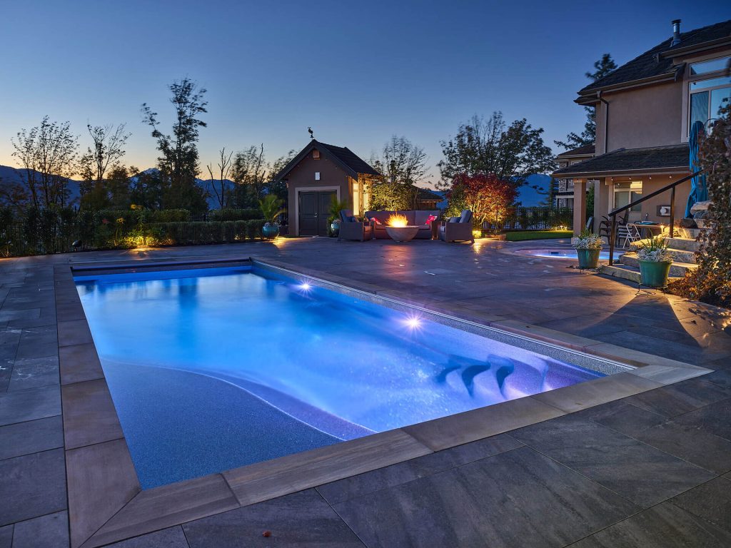 A modern hillside pool illuminated at dusk, featuring underwater LED lighting and a cozy fire-lit seating area.