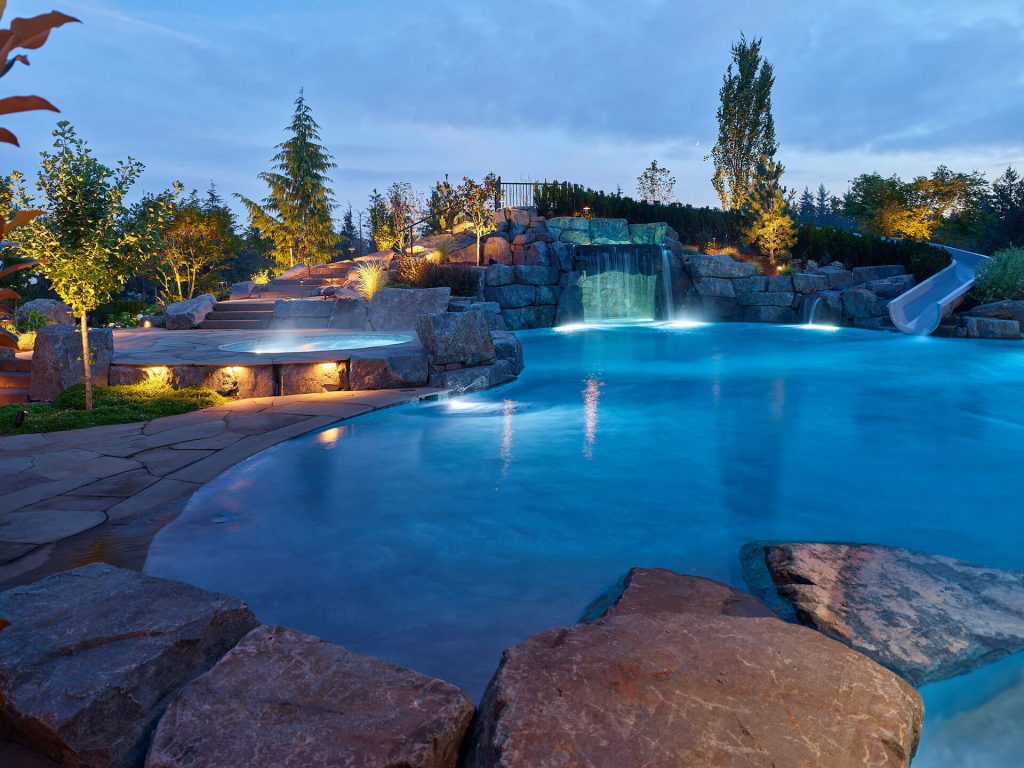 Night view of a resort-style pool featuring illuminated waterfalls, rock accents, and landscaped surroundings.
