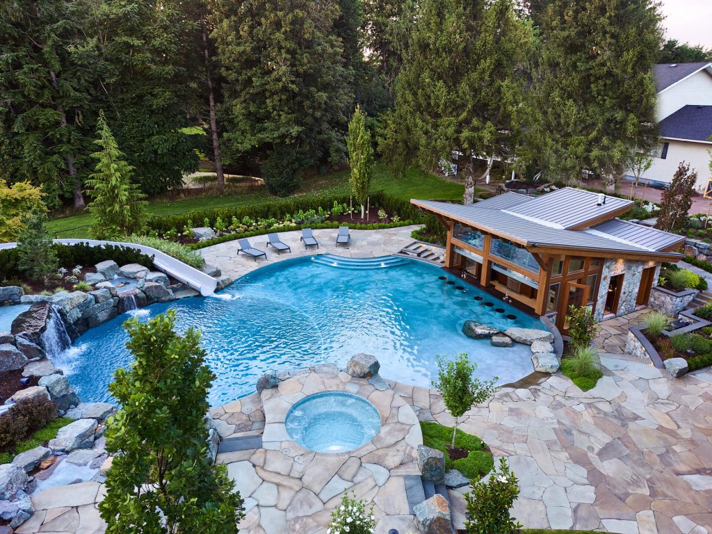 Aerial view of a luxury backyard pool with natural stone decking, spa, and surrounding forest.