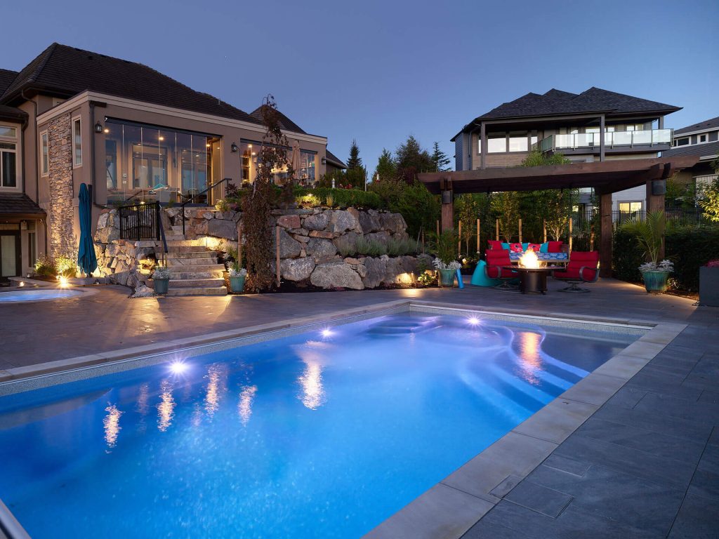 A luxury backyard retreat featuring a lit pool, firepit lounge, and elevated rock terrace.