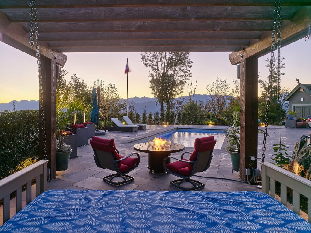 A shaded pergola lounge with firepit seating overlooking a glowing pool and mountain ranges at dusk.