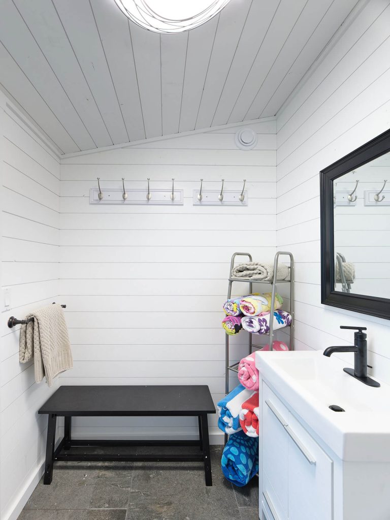 A bright pool house changing room with white shiplap walls, towel hooks, a bench, and modern vanity.