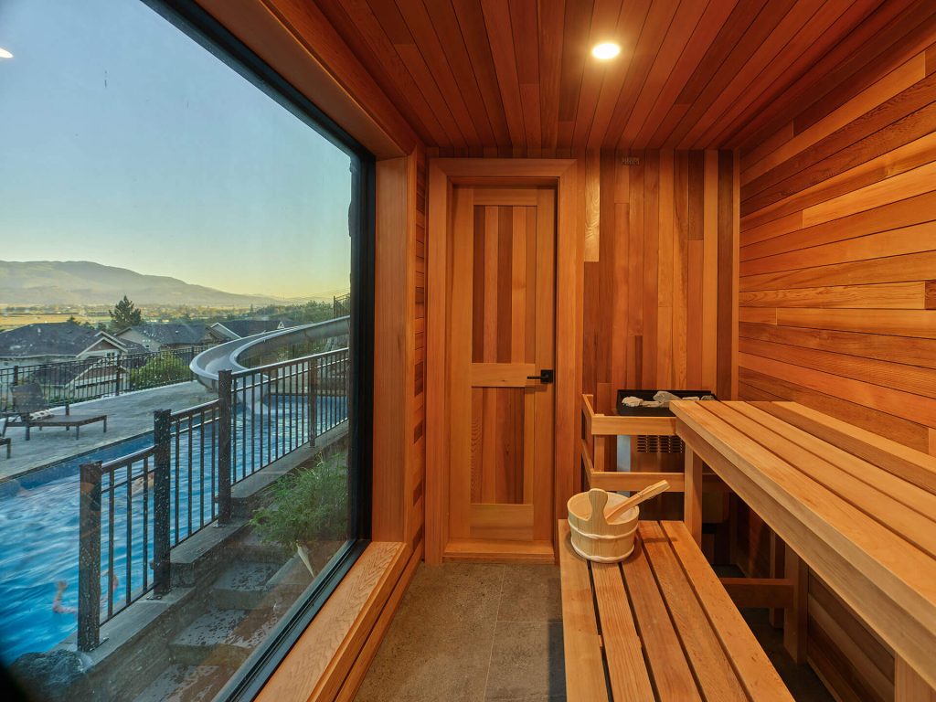 Wood-lined sauna interior with large window overlooking pool and landscape.