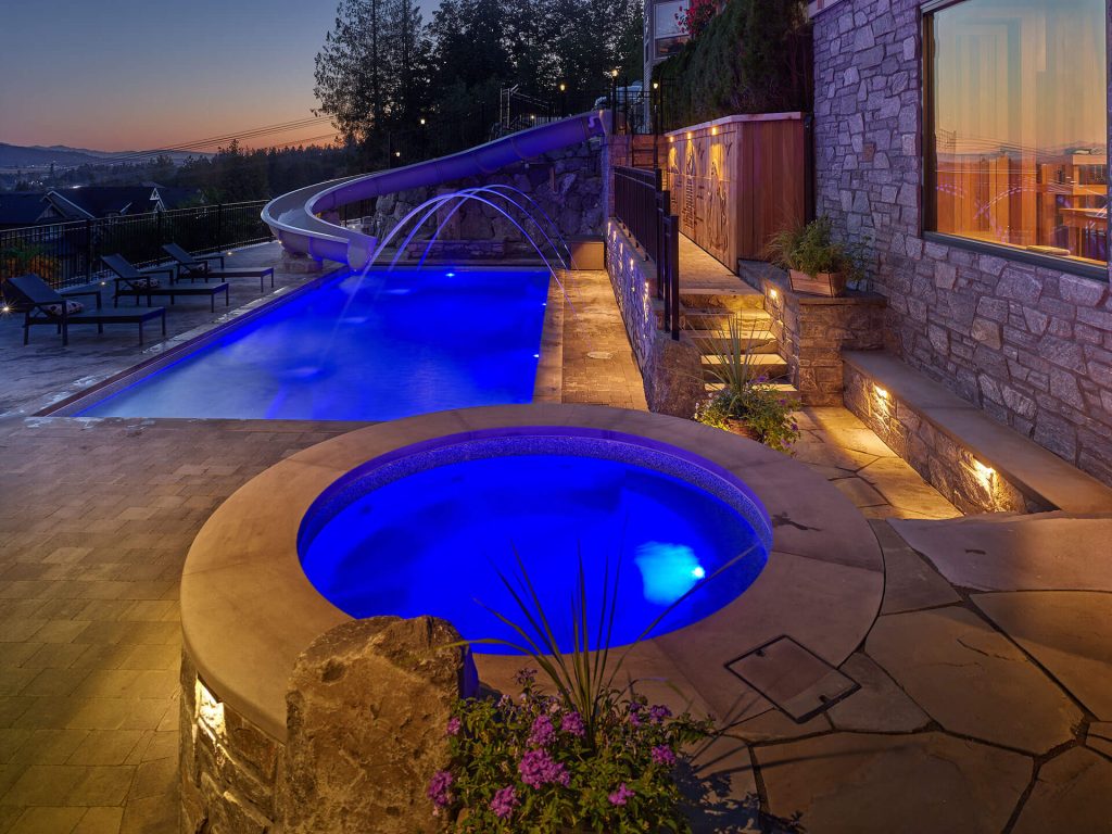 Luxury pool with illuminated waterslide, spa, and stone hardscaping at dusk.