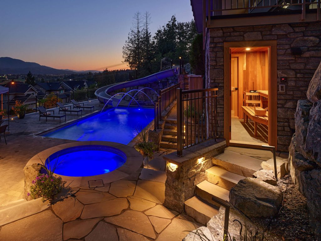 Luxury pool and spa at night with illuminated steps and open sauna doorway.