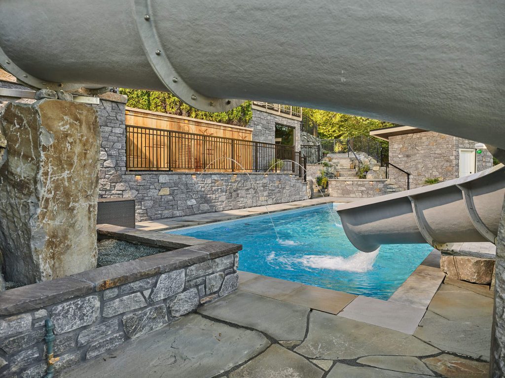 Waterslide over stonework with pool deck jets and surrounding landscaping.