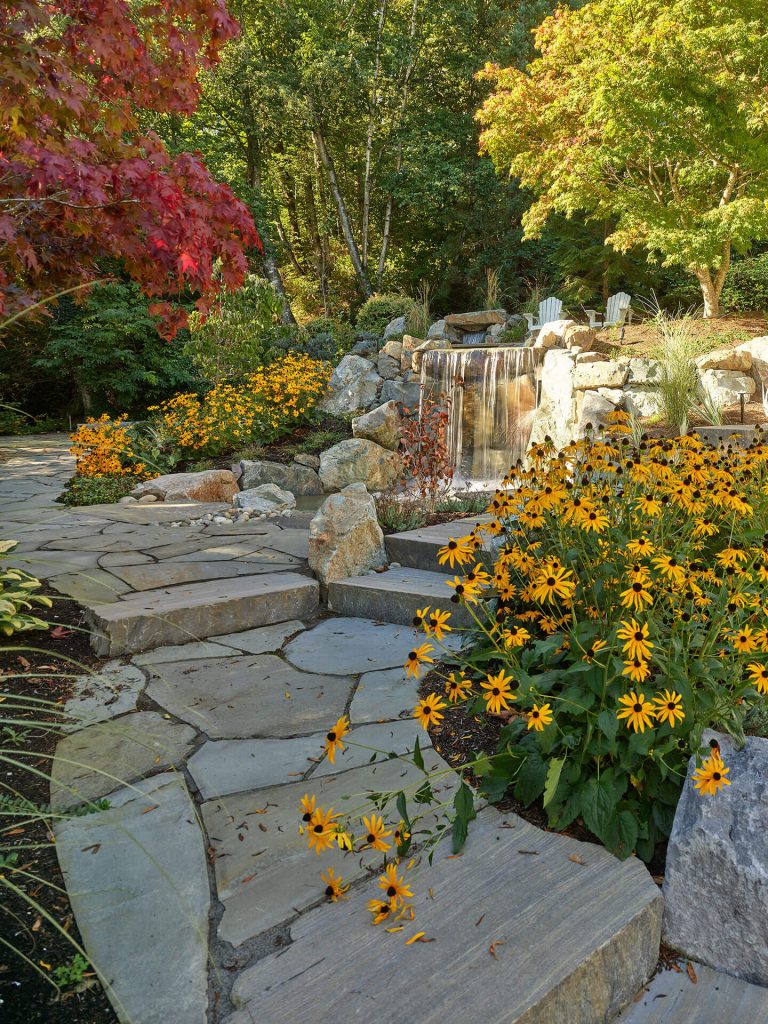 Stone steps curve through blooming yellow flowers toward a rock waterfall in a wooded garden.